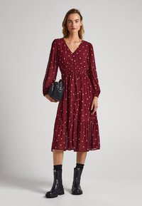 Burgundské šaty s výstřihem do V, dlouhými nafouklými rukávy a smockovaným pasem. Mají vrstvenou sukni se zlatými diamantovými vzory. Kombinované s černými kotníkovými botami.