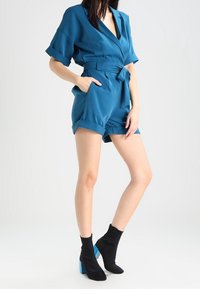 Teal kortärmad jumpsuit med inslagningsdesign, bälte i midjan och uppvikta ärmslut. Kombinerad med svarta ankelskor med blå detaljer.