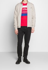 Hombre vistiendo una chaqueta beige con cremallera sobre una camiseta gráfica roja, jeans negros y botas negras con cordones, de pie contra un fondo blanco liso.
