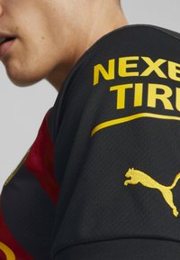 Primer plano de una persona que lleva una camiseta negra y roja de Puma con el texto amarillo "NEXEN TIRE" y el logotipo de Puma en el hombro.