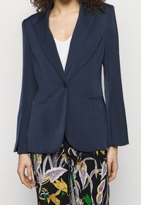 Marinblå skräddarsydd blazer med en knapp, klädda slag och två framfickor. Bärs över en vit topp och mönstrade blommiga byxor.