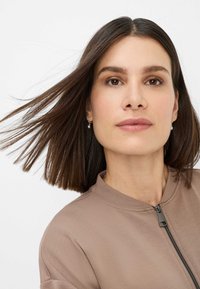Beige zip-up jacket with a rounded collar, smooth texture, and subtle sheen. Model has straight, shoulder-length brown hair and simple earrings.