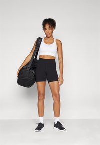 Sorte treningsshorts, hvit sportsbh, sorte joggesko og hvite sokker. Bærer en svart duffelbag med meshinngrep, stående mot en grå bakgrunn.
