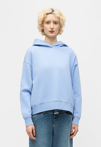 Jeune personne aux cheveux blonds bouclés portant un sweat à capuche bleu clair et un jean bleu, debout devant un fond clair uni.