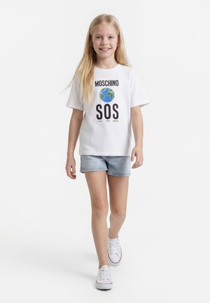 Usmievavé dievča kráčajúce, oblečené v bielom tričku Moschino s grafikou Zeme a nápisom „SOS Save Our Sphere“, svetlé rifľové šortky a biele tenisky.