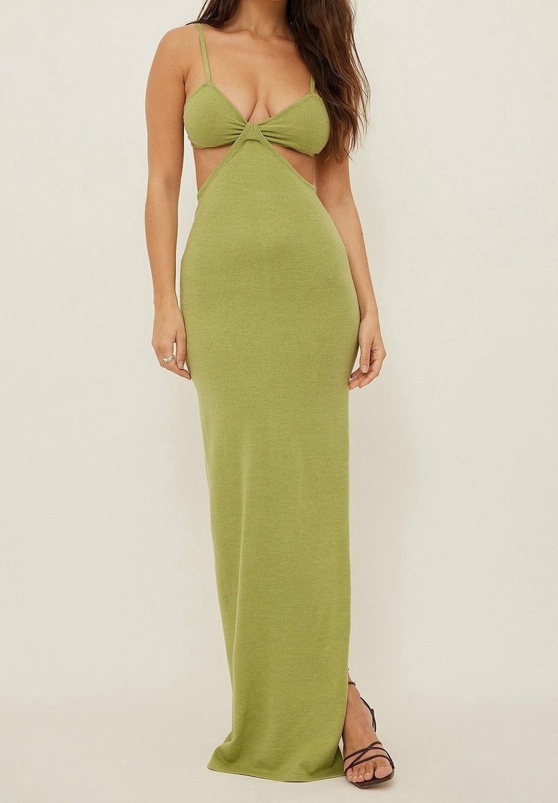 Nainen, joka käyttää oljenväristä, ilman hihansuita maxi-mekkoa ohuilla olkaimilla ja mustia remmi-korkokenkiä, seisoo yksinkertaista taustaa vasten.