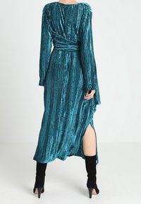 Tyrkysové sametové šaty s dlouhými rukávy a vertikálními pruhy, s překřížením a bočním rozparkem. V kombinaci s vysokými černými botami.