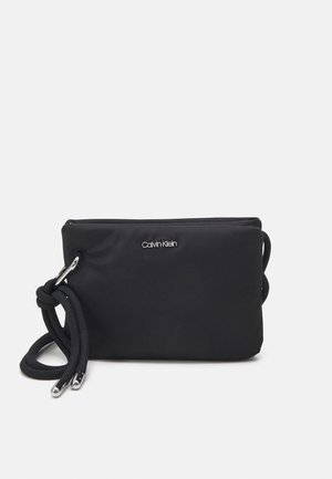 Sac bandoulière rectangulaire Calvin Klein noir avec logo argenté, fermeture éclair en tissu et sangle épaisse tressée avec embouts métalliques sur un fond clair.