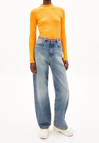 Haut à manches longues côtelé de couleur orange vif, associé à un jean bleu délavé ample, maintenu par une ceinture multicolore, porté avec des baskets blanches.