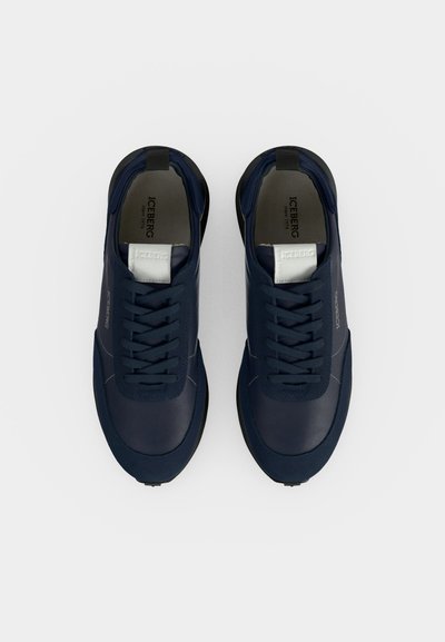Paire de baskets bleu foncé à lacets avec étiquettes blanches sur la languette et semelles noires, vues de dessus sur un fond clair.