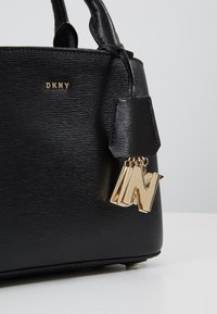 Sac à main en cuir texturé noir avec un logo DKNY doré et un porte-bonheur en forme de lettre 'N'. Poignées courtes, design structuré.