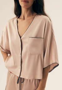 Kobieta nosząca luźną, jasnoróżową satynową bluzkę z czarnym wykończeniem, kieszonką z przodu oraz pasującymi spodniami na sznurek, pokazana od połowy klatki piersiowej do bioder.