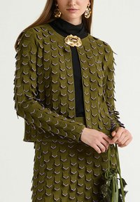 Veste en tricot vert olive avec un design texturé en couches, présentant des accents violets, un fermoir rond et une silhouette ajustée, associée à un haut noir.