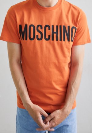 Personne portant un T-shirt orange Moschino et un jean bleu clair, les mains doucement jointes devant un fond neutre.