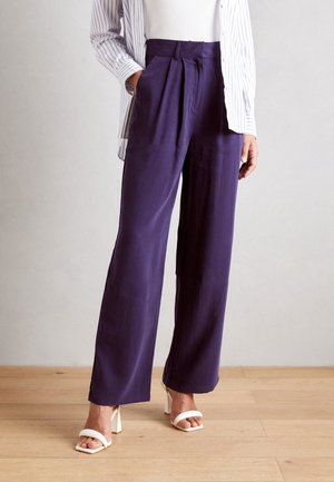 Vrouw met paars, wijduitlopend broek met hoge taille, witte sandalen met hak en een wit topje waar een gestreepte mouw van een overhemd onder zichtbaar is.