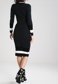 Úzké černé šaty s bílými pruhovanými detaily, s kulatým výstřihem a dlouhými rukávy. Nosené s černými kotníkovými botami s květinovou výšivkou.