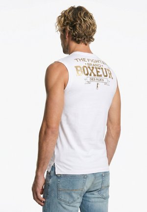 Mężczyzna o kręconych blond włosach nosi białą koszulkę bez rękawów z złotym napisem „THE FIGHTER BRAND BOXEUR DES RUES” oraz jasnoniebieskie dżinsy, stoi tyłem.
