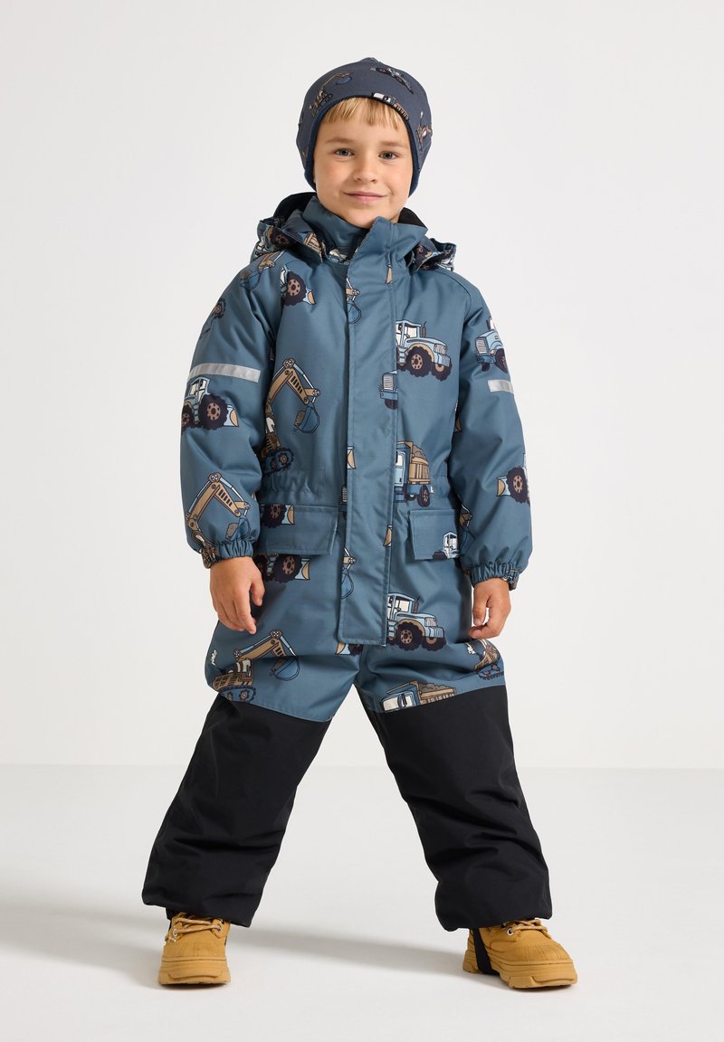 Veste imperméable bleue et salopette avec des motifs de tracteurs, accents noirs et détails réfléchissants, assorties d'un chapeau bleu foncé et de bottes jaunes.