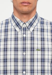 Blå og hvit rutete skjorte med krage som kan kneppes ned, en blanding av store og små rutemønstre, med en grønn brodert logo på brystet.