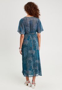 Robe maxi à motifs bleus avec des manches courtes en dentelle et une taille élastique. Elle présente une fente latérale et un tissu fluide et léger. Sandales argentées.