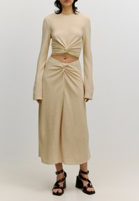 Beige Zweiteiler aus strukturiertem Stoff; kurzes Oberteil mit langen Ärmeln und Knotendetail sowie einen Rock mit mittlerer Taille, der ein ähnliches Drehdesign aufweist. Braune Blockabsatzsandalen.