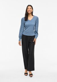 Blauwe gebreide trui met V-hals en gestructureerde patronen, gecombineerd met zwarte wijde broek en zwarte open-toe hakken.