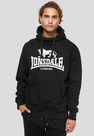 Jeune homme aux cheveux bouclés portant un sweat à capuche noir Lonsdale London avec un logo blanc et une poche kangourou, mains partiellement dans les poches, fond neutre.