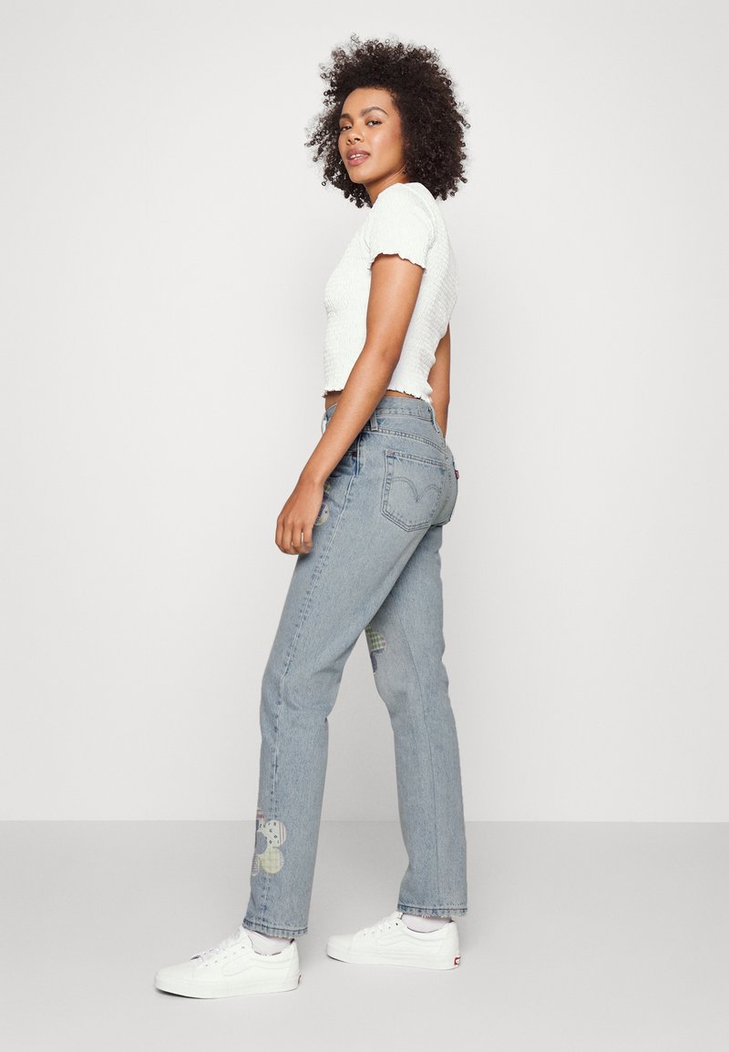 Jeans de mezclilla de color azul claro con diseños de flores en patchwork en las piernas inferiores, combinados con una camiseta corta texturizada blanca y zapatillas blancas.