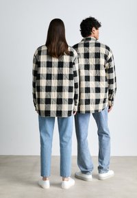 Juostuotas tarkuotas marškiniai juodos ir kreminės spalvos, su ilgomis rankovėmis ir užsegami rankogaliai, derinami su šviesiai mėlynomis džinsais ir baltomis sportbačiais.