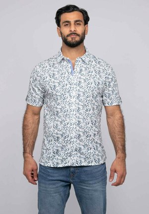 Homme barbu portant une chemise blanche à manches courtes avec un motif de feuilles bleues et un jean bleu, debout devant un fond uni.