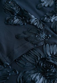 Melna teksturēta auduma ar paaugstinātu ziedu izšuvumu ar detalizētām ziedlapiņām un spirālēm. Dizains ir sarežģīts, ar dažādām formām un izmēriem.