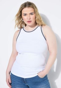 Witte mouwloze top met een geribbeld textuur, voorzien van een zwart-wit bloemenrand langs de halslijn en armsgaten. Gecombineerd met blauwe jeans.