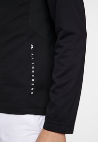 Svart atletisk långärmad topp med textilstruktur, med en sidologotyp i vitt och en figurnära design. Ärmarna är släta.