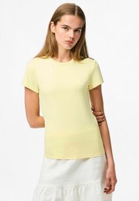 Korte mouwen, ronde hals gele t-shirt gemaakt van een gladde stof. Aansluitend ontwerp, gedragen met een witte gelaagde rok, tegen een lichte achtergrond.