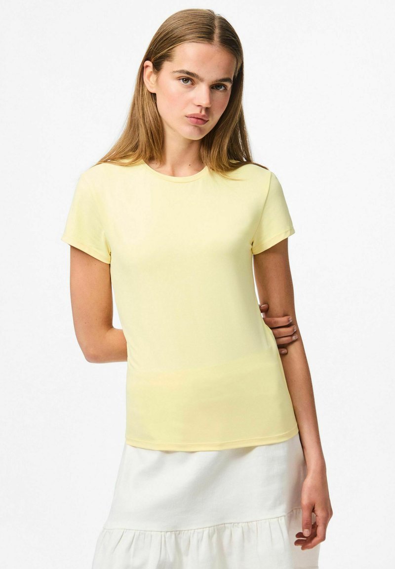 Korte mouwen, ronde hals gele t-shirt gemaakt van een gladde stof. Aansluitend ontwerp, gedragen met een witte gelaagde rok, tegen een lichte achtergrond.