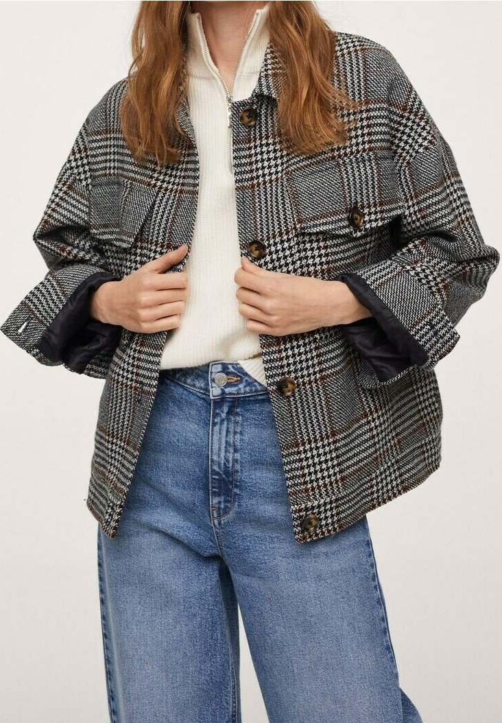 Personne portant une veste à carreaux noir et blanc boutonnée sur un pull zippé blanc et un jean bleu taille haute, les mains ajustant le devant de la veste.