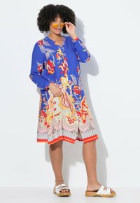 Blaues Kleid mit lebhaften floralen Mustern in Rot, Gelb und Pink; lange Ärmel; Knopfleiste vorne; mittlere Oberschenkellänge; kombiniert mit weißen Sandalen.