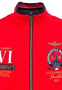 Giacca bomber rossa con colletto alto, chiusura con zip, logo ricamato e dettagli decorativi in oro e bianco.
