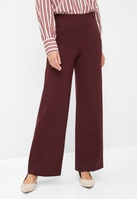 Pantaloni a gamba larga bordeaux realizzati in tessuto liscio, con vita alta e dettagli in cucitura. Abbinati a una blusa a maniche lunghe a righe e ballerine.