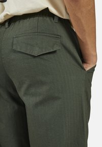 Green herringbone pants with an elastic waistband, side pocket, and a single back pocket featuring a diamond flap. Textured fabric.