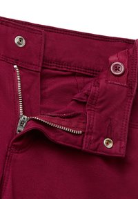 Acercamiento de la cremallera abierta en unos pantalones burdeos, con botón visible y cierre de presión metálico en la cintura.