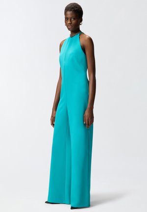 Donna in piedi, indossa una tuta senza maniche color teal con pantaloni ampi e collo alto, accessoriata con una collana a catena e orecchini.