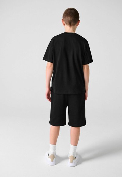 Garçon de dos portant un t-shirt noir, un short noir, des chaussettes blanches et des baskets blanches sur un fond clair uni.