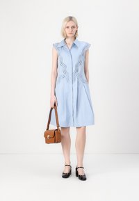 Abito camicia senza maniche di colore azzurro chiaro, con design scallop e due tasche anteriori. Abbinato a una piccola borsa a tracolla marrone e scarpe nere.