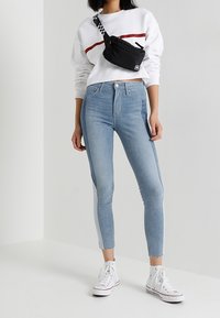 Woman wearing cropped white sweatshirt with red stripe, two-tone skinny jeans, white high-top sneakers, and black crossbody bag with checkered strap.