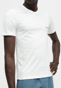 Homme portant un t-shirt blanc uni col en V à manches courtes avec un petit logo rouge "HUGO" sur la poitrine, associé à un pantalon bleu texturé.