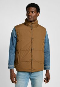 Brun, vadderad väst med hög krage, som har tryckknappar och kontrasterande ärmar i blå denim. Lätt och vattentåligt material.