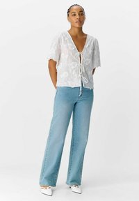 Hvid blomstret broderet top med korte ærmer, bundet foran, parret med lyseblå høj talje jeans og hvide spidse sko.