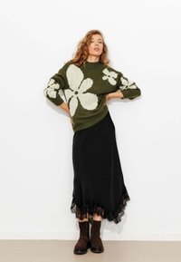 Pull en maille vert olive avec un motif floral blanc, associé à une jupe noire avec des bordures en dentelle et des bottines brunes.