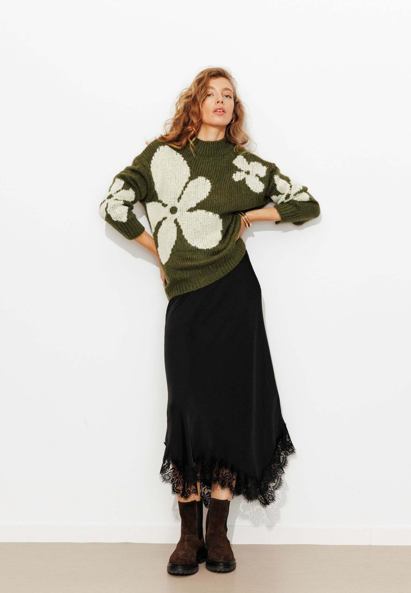 Pull en maille vert olive avec un motif floral blanc, associé à une jupe noire avec des bordures en dentelle et des bottines brunes.